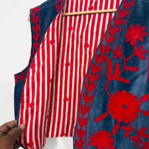 May include: A sleeveless vest featuring a deep blue exterior with vibrant red floral embroidery along the front edges. The interior lining is made of a white and red vertical striped fabric with small red heart shapes interspersed throughout.