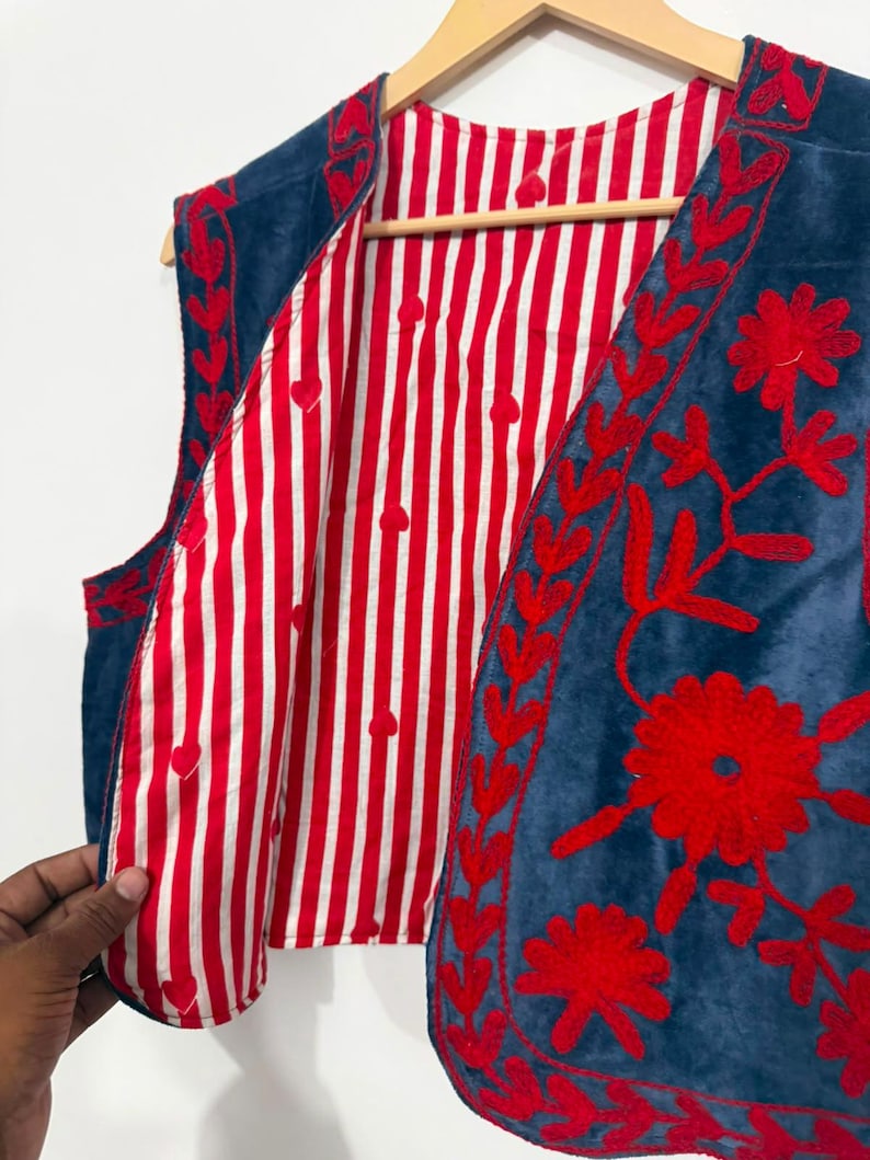 May include: A sleeveless vest featuring a deep blue exterior with vibrant red floral embroidery along the front edges. The interior lining is made of a white and red vertical striped fabric with small red heart shapes interspersed throughout.