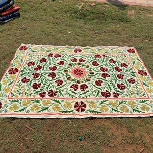 May include: A rectangular, cream-colored embroidered textile with a floral design. The design features red and maroon flowers, green leaves, and yellow accents. The border is a light pink color. A stack of folded textiles is in the background.