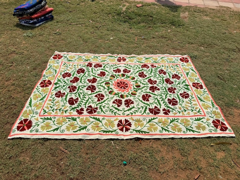 May include: A rectangular, cream-colored embroidered textile with a floral design. The design features red and maroon flowers, green leaves, and yellow accents. The border is a light pink color. A stack of folded textiles is in the background.