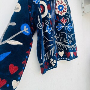 May include: A navy blue jacket with intricate floral embroidery in red, white, and blue. The jacket features a velvet texture and a detailed design of flowers, hearts, and swirling patterns. The sleeve is visible, showcasing the elaborate embroidery.