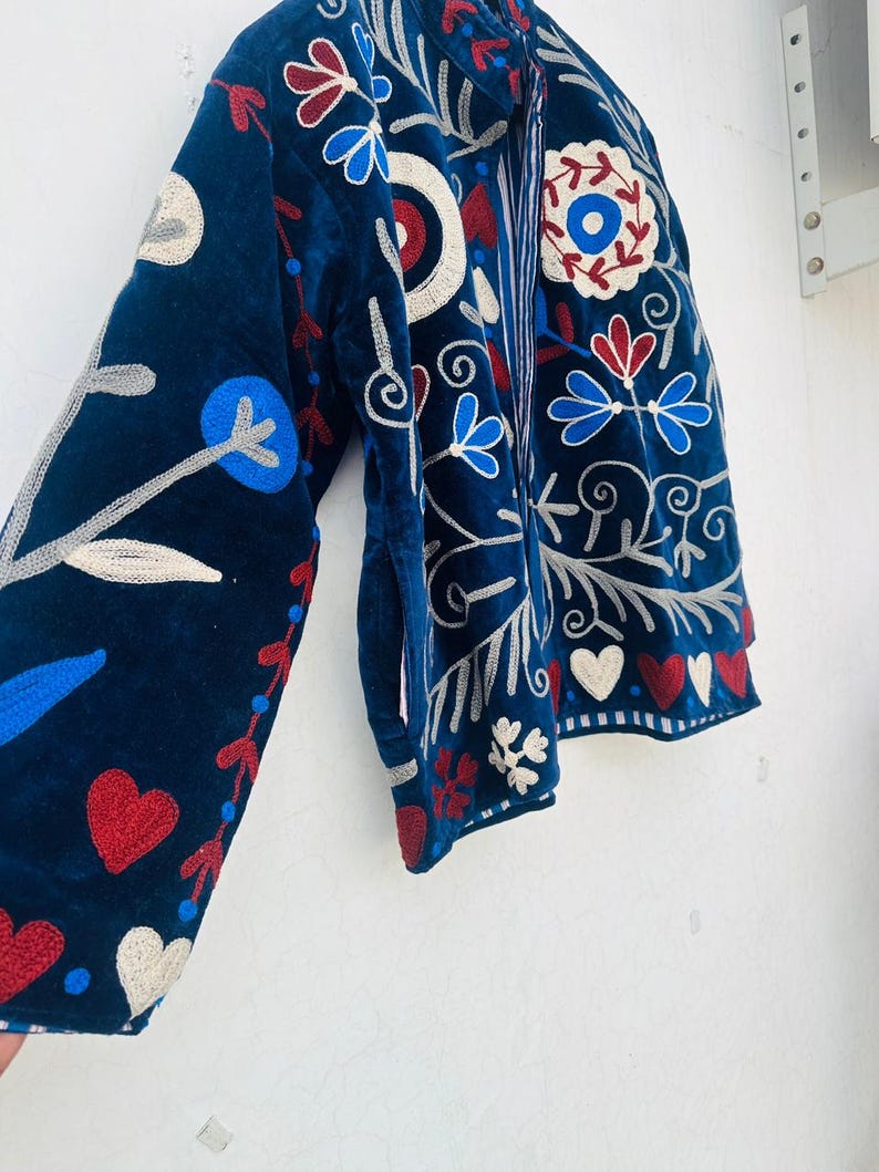May include: A navy blue jacket with intricate floral embroidery in red, white, and blue. The jacket features a velvet texture and a detailed design of flowers, hearts, and swirling patterns. The sleeve is visible, showcasing the elaborate embroidery.
