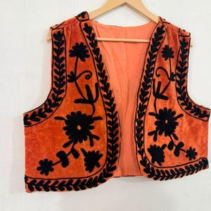 K&ouml;nnte beinhalten: Eine &auml;rmellose Weste in einem leuchtenden Orangeton mit aufwendigen schwarzen Blumenstickereien entlang der Vorderteile und Kanten. Das Kleidungsst&uuml;ck ist vorne offen und hat ein einfarbiges orangefarbenes Innenfutter mit einem detaillierten Blumenmuster.