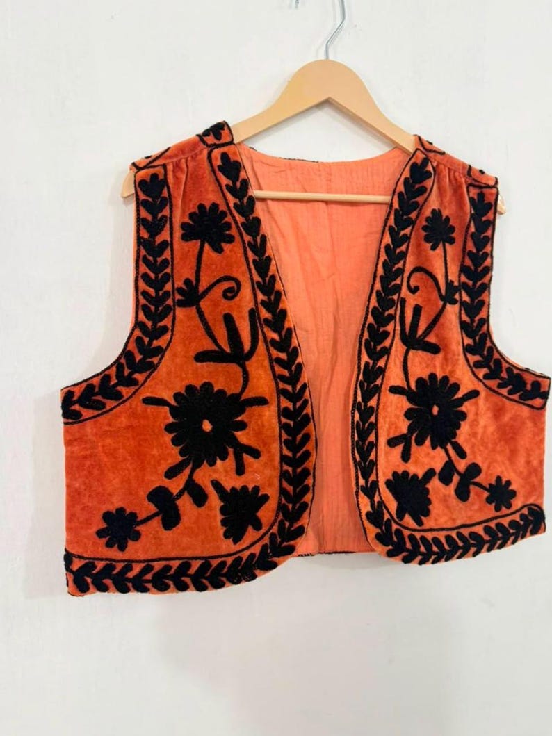 K&ouml;nnte beinhalten: Eine &auml;rmellose Weste in einem leuchtenden Orangeton mit aufwendigen schwarzen Blumenstickereien entlang der Vorderteile und Kanten. Das Kleidungsst&uuml;ck ist vorne offen und hat ein einfarbiges orangefarbenes Innenfutter mit einem detaillierten Blumenmuster.