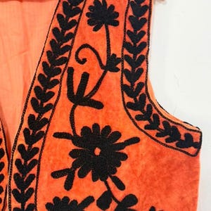 K&ouml;nnte beinhalten: Eine &auml;rmellose orangefarbene Weste mit schwarzer Blumen- und Blattstickerei entlang der Kanten und Vorderteile. Das Kleidungsst&uuml;ck besteht aus einem weich strukturierten Stoff und wird auf einem Holzkleiderb&uuml;gel vor einem schlichten Hintergrund pr&auml;sentiert.