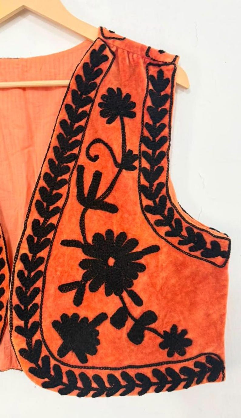 K&ouml;nnte beinhalten: Eine &auml;rmellose orangefarbene Weste mit schwarzer Blumen- und Blattstickerei entlang der Kanten und Vorderteile. Das Kleidungsst&uuml;ck besteht aus einem weich strukturierten Stoff und wird auf einem Holzkleiderb&uuml;gel vor einem schlichten Hintergrund pr&auml;sentiert.