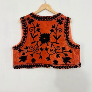 K&ouml;nnte beinhalten: Eine &auml;rmellose Weste in einem leuchtenden gebrannten Orangeton mit aufwendiger schwarzer Blumenstickerei auf der Vorderseite. Das Design umfasst eine detaillierte Bord&uuml;re entlang des Ausschnitts, der Armausschnitte und des Saums.