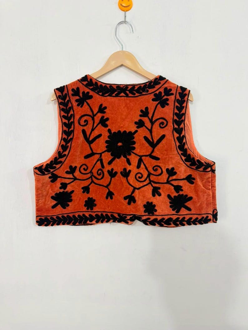 K&ouml;nnte beinhalten: Eine &auml;rmellose Weste in einem leuchtenden gebrannten Orangeton mit aufwendiger schwarzer Blumenstickerei auf der Vorderseite. Das Design umfasst eine detaillierte Bord&uuml;re entlang des Ausschnitts, der Armausschnitte und des Saums.