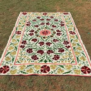 May include: A large, rectangular, off-white textile with an intricate floral design. The pattern features burgundy, green, and yellow flowers with a coral border. The textile is laid out on a grassy surface.