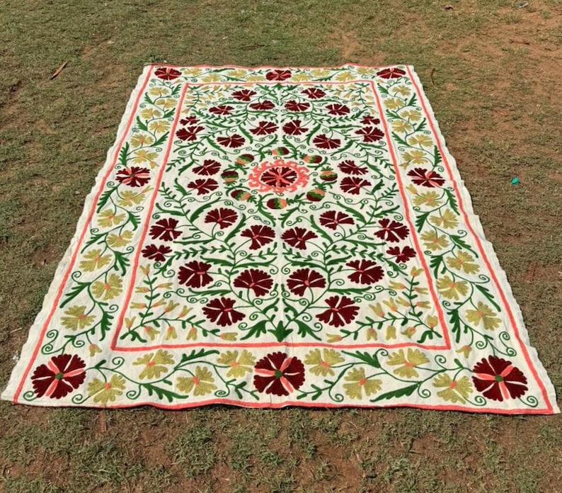 May include: A large, rectangular, off-white textile with an intricate floral design. The pattern features burgundy, green, and yellow flowers with a coral border. The textile is laid out on a grassy surface.