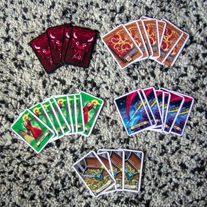 Puede incluir: Una colección de cartas de juego con varias ilustraciones. Las cartas están dispuestas en cuatro grupos, cada uno con un tema diferente. El primer grupo presenta cartas rojas con una ilustración de un toro. El segundo grupo presenta cartas verdes con una mujer sosteniendo un bastón. El tercer grupo presenta cartas azules con una ilustración de un rayo. El cuarto grupo presenta cartas blancas con un cofre lleno de monedas de oro.