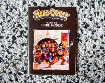 Può includere: Un libro marrone intitolato "Hero Quest Against the Ogre Horde" con un'illustrazione colorata di personaggi fantasy. Il libro è ambientato su uno sfondo a motivi bianchi e neri.