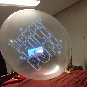 May include: A large, clear balloon with the text "BLOW ME TILL I POP!" in a blue, outlined font. The balloon is decorated with blue star shapes. The balloon is inflated and appears to be indoors, resting on a red bedspread.