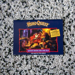 May include: A blue and red book cover with the title "HeroQuest" and the text "Quest Book for the Elf: The Mage of the Mirror". The cover features an illustration of a fantasy scene with characters in a dark room.