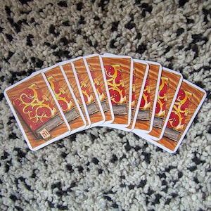 Puede incluir: Un juego de 10 cartas de juego con un diseño de libro rojo y dorado. Las cartas están dispuestas en forma de abanico, con el dorso de las cartas mirando al espectador.