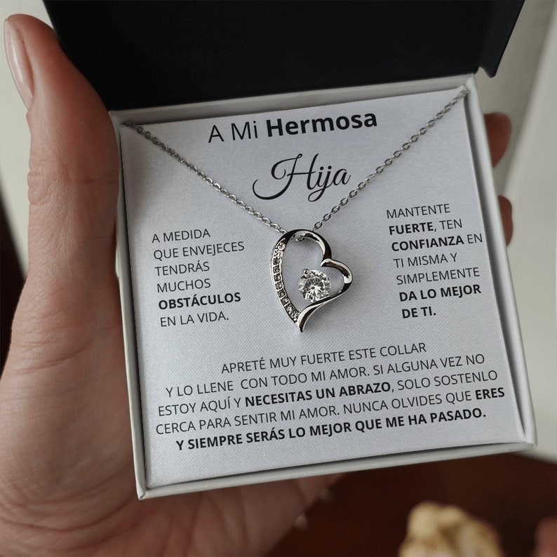 May include: A silver necklace with a heart pendant featuring a sparkling crystal. The necklace comes in a white box with a message that reads "A Mi Hermosa Hija" and includes a heartfelt message about strength, confidence, and love.