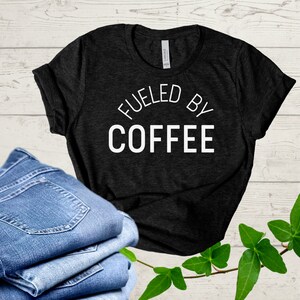 Pode incluir: Uma camiseta preta com o texto "Fueled by Coffee" em letras brancas na frente. A camiseta está sobre uma superfície de madeira com um par de calças de ganga azuis e folhas verdes.