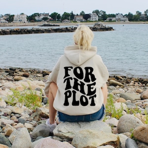 May include: A person wearing a cream-colored hoodie with the text "FOR THE PLOT" in black lettering on the back. The person is sitting on rocks by the water.