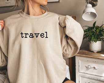 Könnte beinhalten: Ein beiger Pullover mit dem Wort "travel" in Schwarz auf der Vorderseite.
