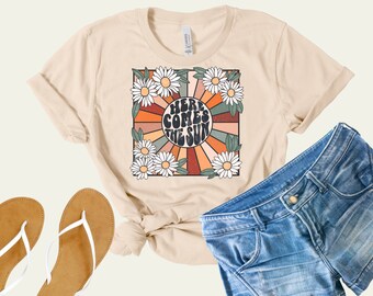 May include: A beige t-shirt with a retro-style graphic featuring a sunburst design with daisies and the text "Here Comes The Sun".