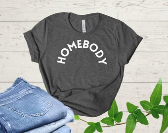 Puede incluir: Una camiseta gris oscuro con el texto "HOMEBODY" impreso en blanco en forma de arco.