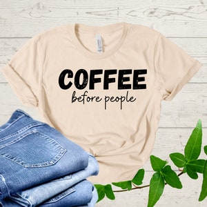 Puede incluir: Una camiseta beige con texto negro que dice "COFFEE before people". La camiseta está doblada y se muestra sobre una superficie de madera con pantalones vaqueros azules y hojas verdes.