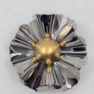 May include: A silver flower brooch with a gold center. The flower has a ruffled design and the center has a round, raised gold button.
