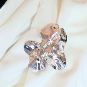 May include: A silver-coloured metal brooch with an abstract, organic design. The brooch has a fluid, rippled texture, reflecting light and creating a dynamic appearance. The brooch is set against a backdrop of soft, white fabric.