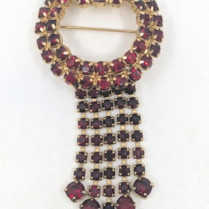 May include: A gold tone brooch with a circle of red rhinestones and a dangling fringe of red rhinestones.