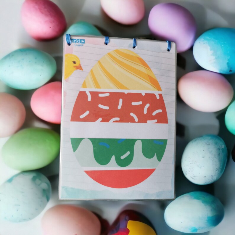 K&ouml;nnte beinhalten: Ein Notizbuch mit einem bunten Osterei-Design auf dem Cover. Das Ei ist mit roten, gr&uuml;nen und gelben Streifen und Streuseln verziert. Das Notizbuch ist von pastellfarbenen Ostereiern umgeben.