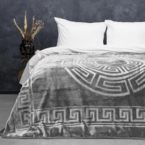 May include: A gray blanket with a geometric pattern draped over a bed with white pillows. The blanket features a large, circular design in a lighter shade of gray, with a border of repeating square shapes. The bed is set against a dark gray wall.