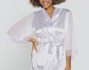May include: A white satin robe with sheer sleeves and lace trim. The robe has a tie waist and a V-neckline. The model is smiling and has curly hair. The robe is a light, silky material.