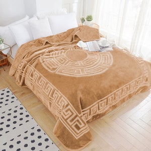May include: A tan-colored blanket with a Greek key pattern and a circular design in the center. The blanket is draped over a bed with white pillows. The blanket is made of a soft, plush material.