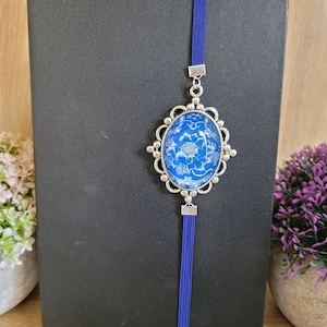 May include: A blue and white floral design bookmark with a silver metal frame and a blue tassel. The bookmark is attached to a black notebook.