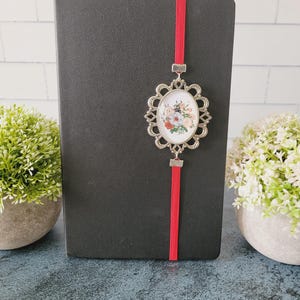 May include: A black notebook featuring a vertical red elastic band closure. Attached to the band is an oval silver-toned metal pendant with a decorative scalloped edge, framing a small, colorful floral illustration of roses and greenery.