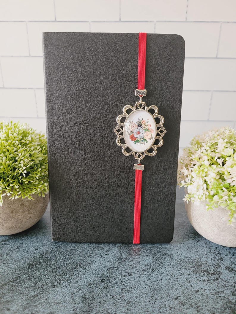 May include: A black notebook featuring a vertical red elastic band closure. Attached to the band is an oval silver-toned metal pendant with a decorative scalloped edge, framing a small, colorful floral illustration of roses and greenery.