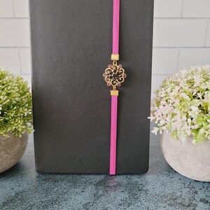 May include: A black notebook secured with a vertical hot pink elastic band. Centered on the band is a circular gold-toned metal ornament featuring intricate filigree scrollwork and small dark purple crystals arranged in a floral pattern.