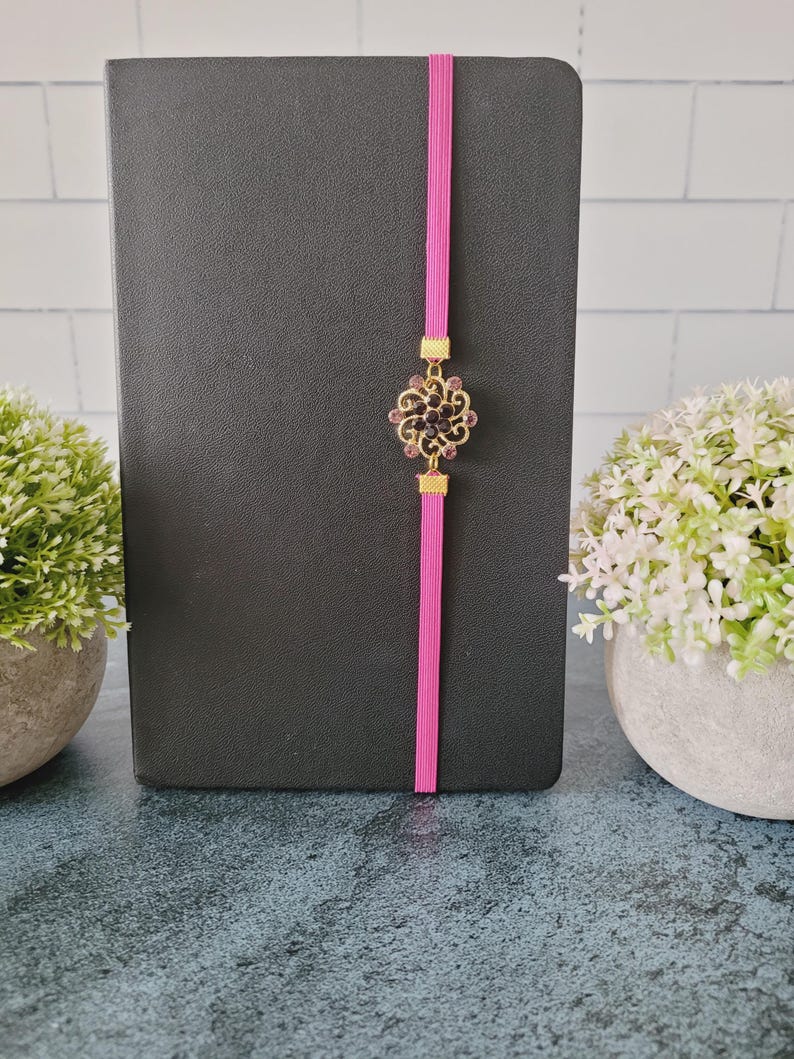 May include: A black notebook secured with a vertical hot pink elastic band. Centered on the band is a circular gold-toned metal ornament featuring intricate filigree scrollwork and small dark purple crystals arranged in a floral pattern.
