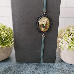 May include: A black notebook featuring a vertical teal elastic band closure. Attached to the band is an oval-shaped black metal frame containing a miniature print of a bouquet of light pink roses in a green vase against a dark background.