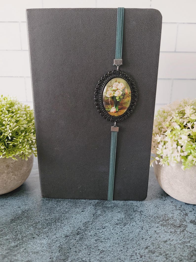 May include: A black notebook featuring a vertical teal elastic band closure. Attached to the band is an oval-shaped black metal frame containing a miniature print of a bouquet of light pink roses in a green vase against a dark background.