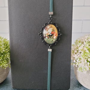 May include: A black notebook featuring a vertical teal elastic band. Attached to the band is an oval-shaped black metal pendant with a decorative scalloped edge, framing a colorful floral still life painting with red, yellow, and white flowers.