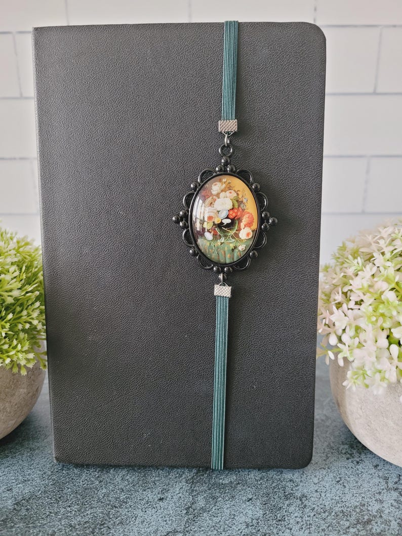 May include: A black notebook featuring a vertical teal elastic band. Attached to the band is an oval-shaped black metal pendant with a decorative scalloped edge, framing a colorful floral still life painting with red, yellow, and white flowers.