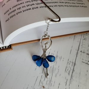 May include: A silver bookmark with a blue ballerina charm. The ballerina is wearing a blue skirt and has a silver crown on her head.