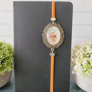 May include: A black notebook featuring an orange elastic band closure with an attached oval-shaped antique bronze-toned metal pendant. The pendant contains a white insert decorated with a colorful floral illustration of orange, pink, and yellow flowers.