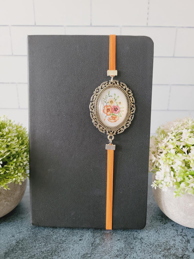 May include: A black notebook featuring an orange elastic band closure with an attached oval-shaped antique bronze-toned metal pendant. The pendant contains a white insert decorated with a colorful floral illustration of orange, pink, and yellow flowers.