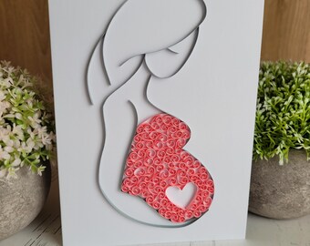 May include: A white greeting card featuring a paper quilling art design of a pregnant figure. The silhouette is outlined in thin grey paper strips, while the belly area is filled with numerous small, coiled pink paper rolls and a heart shape.