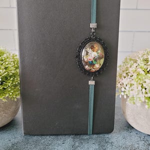 May include: A dark gray notebook featuring a teal elastic band closure. Attached to the band is an oval-shaped black filigree pendant containing a miniature floral still life painting with pink, white, and yellow flowers in a blue vase.