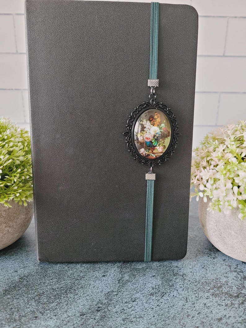 May include: A dark gray notebook featuring a teal elastic band closure. Attached to the band is an oval-shaped black filigree pendant containing a miniature floral still life painting with pink, white, and yellow flowers in a blue vase.
