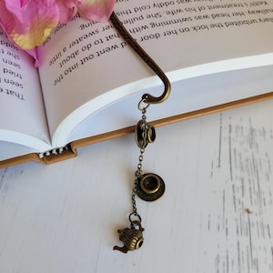 May include: A bronze bookmark with a chain and charms shaped like a teapot, saucer, and teacup. The bookmark is placed in an open book with a pink flower on the top of the page.