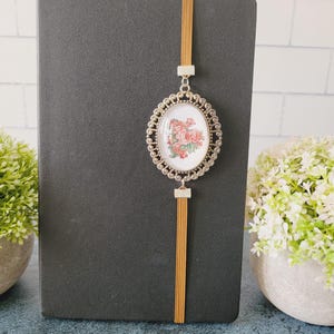 May include: A black notebook featuring a vertical mustard-colored elastic band. Attached to the band is an oval-shaped silver-toned metal pendant with a decorative filigree border, framing a white insert with a pink and green floral illustration.